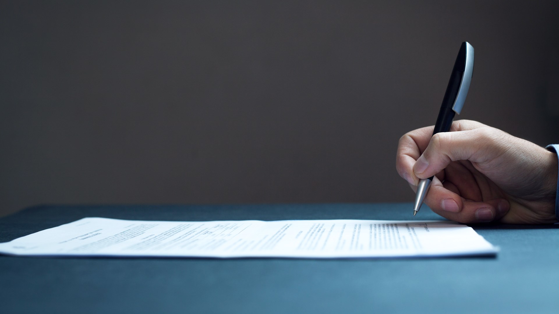 A hand holding a pen, poised to sign a printed agreement on a dark desk — the signature that turns a document into a decision.
